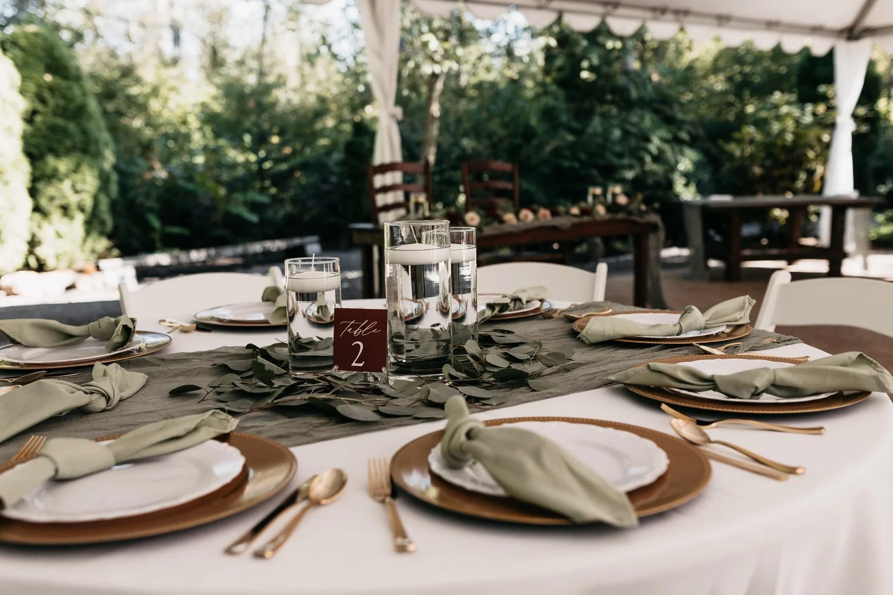 A reception table under the tent set with gold charger plates, brushed-gold flatware, and a sage-green foliage runner