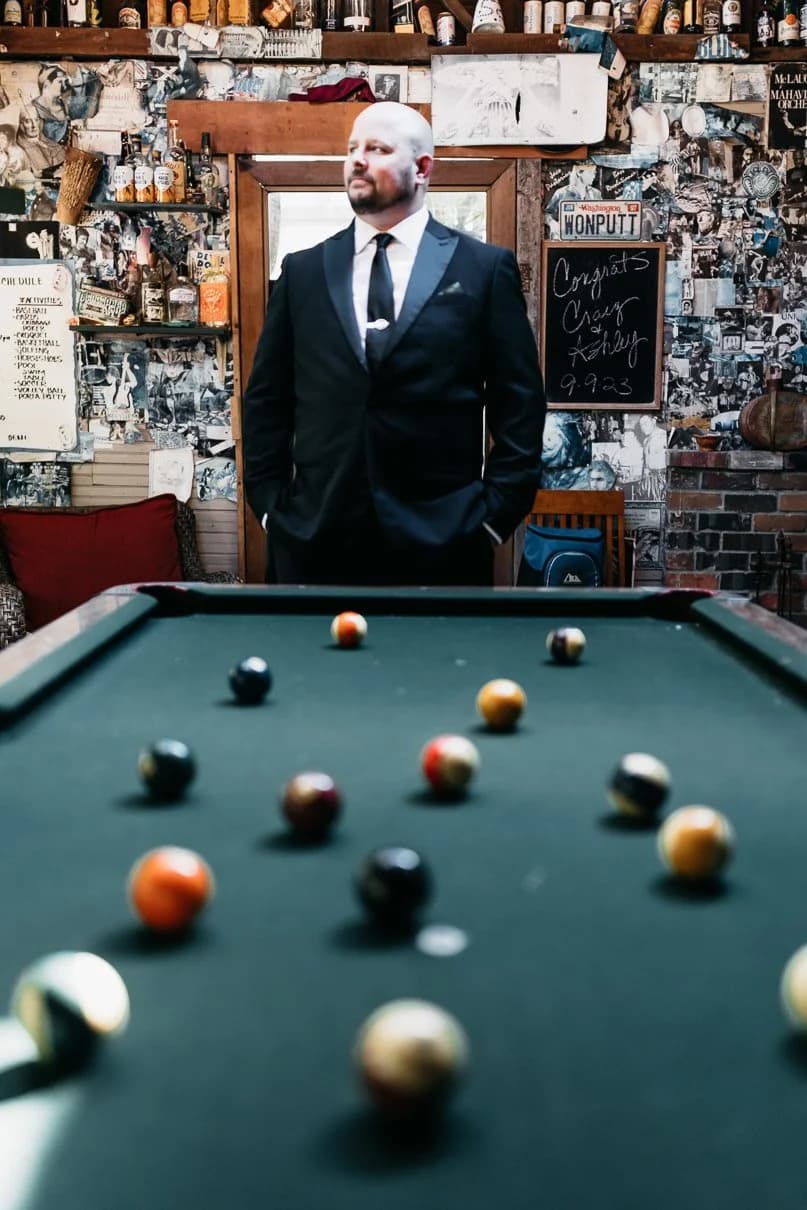 The groom standing in the Pub behind the pool table, the walls behind him layered with framed photographs and memorabilia