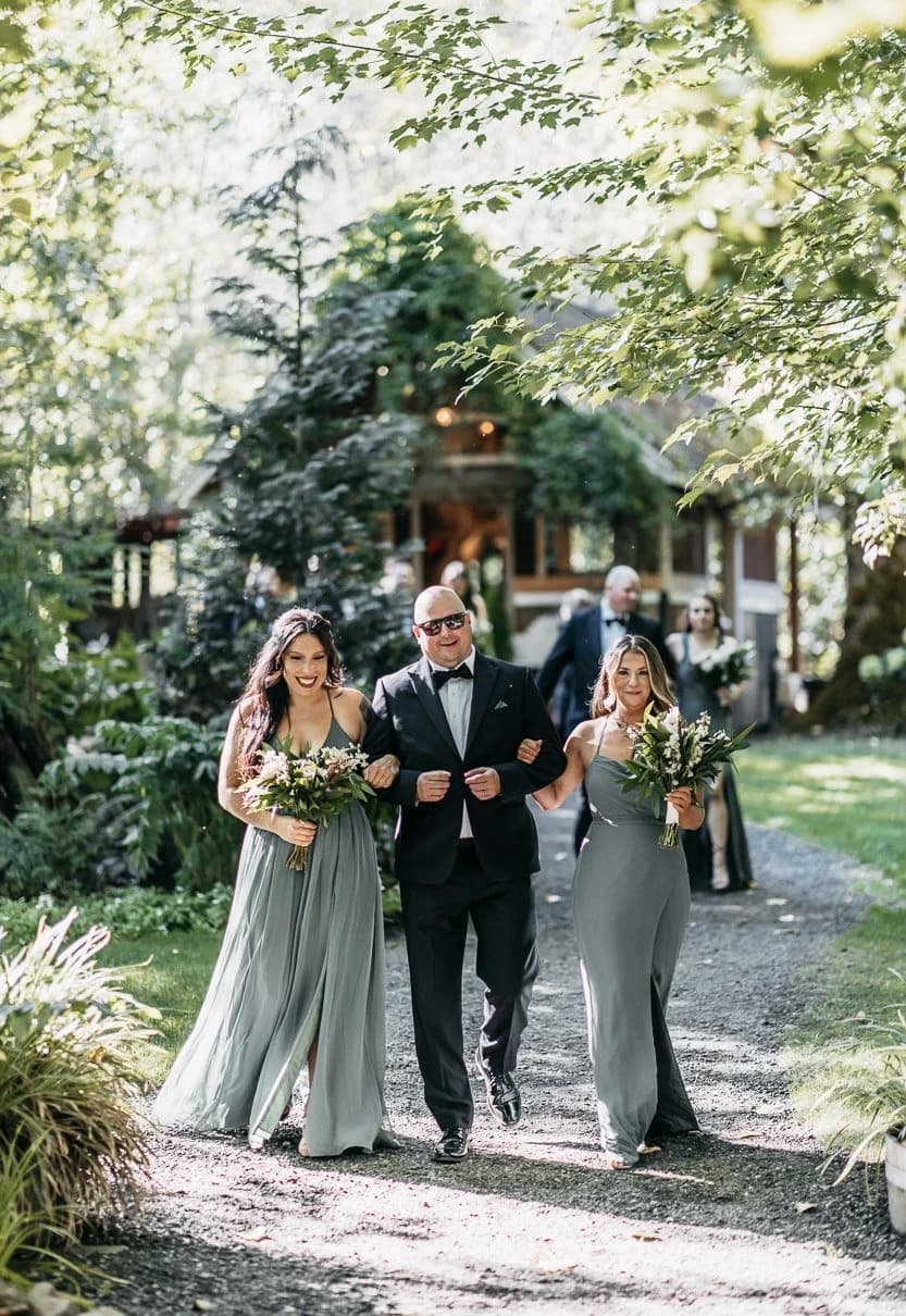 A wedding party walking a garden path between tall firs toward the house, bouquets in hand
