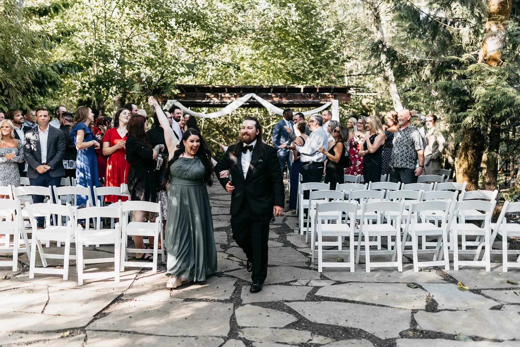 A newly married couple walking back down the stone aisle lined with white chairs beneath the fir canopy, guests cheering on both sides